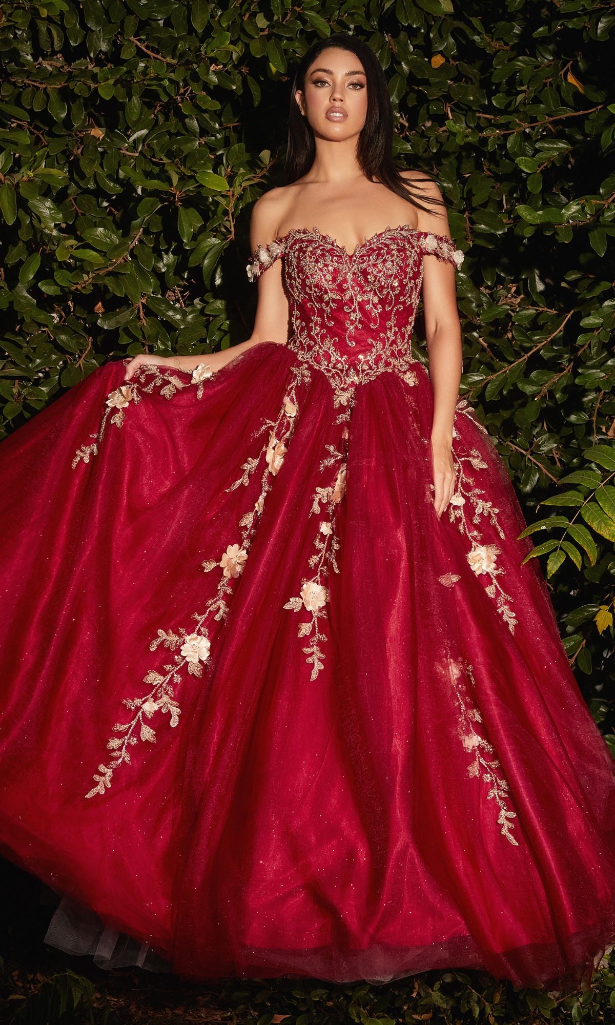 A woman stands gracefully before lush green foliage, wearing the Off-Shoulder Long Sweetheart Prom Ball Gown CD0185 in deep red with gold floral-lace appliques.